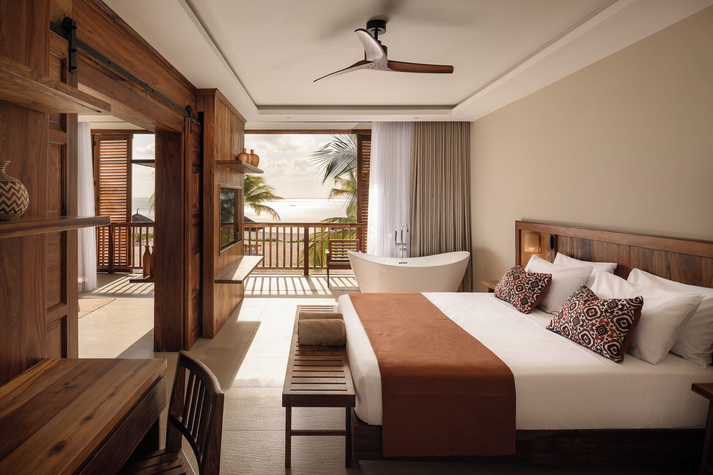 Hotelkamer met een bed, een houten bureau, een bankje, een plafondventilator en uitzicht op het strand.