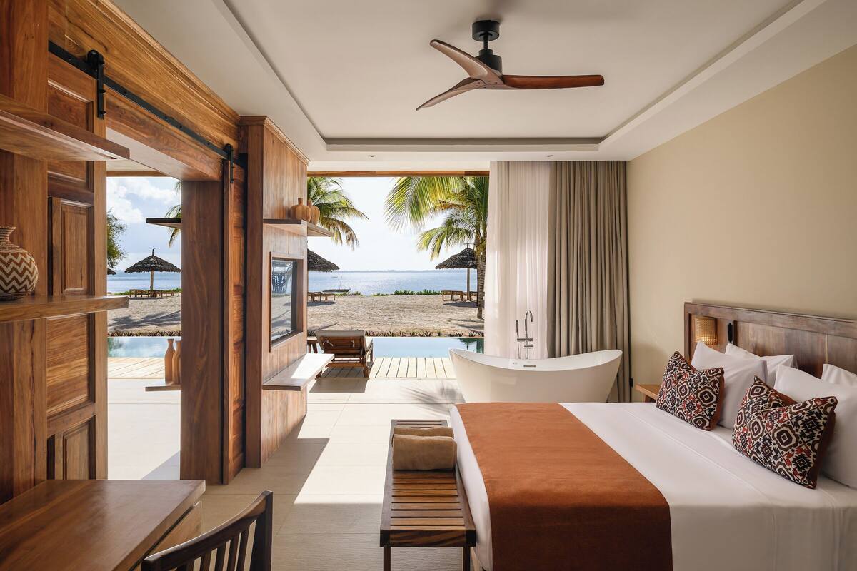 A hotel room with a bed, a bathtub, a dining area, and a view of the beach and ocean.