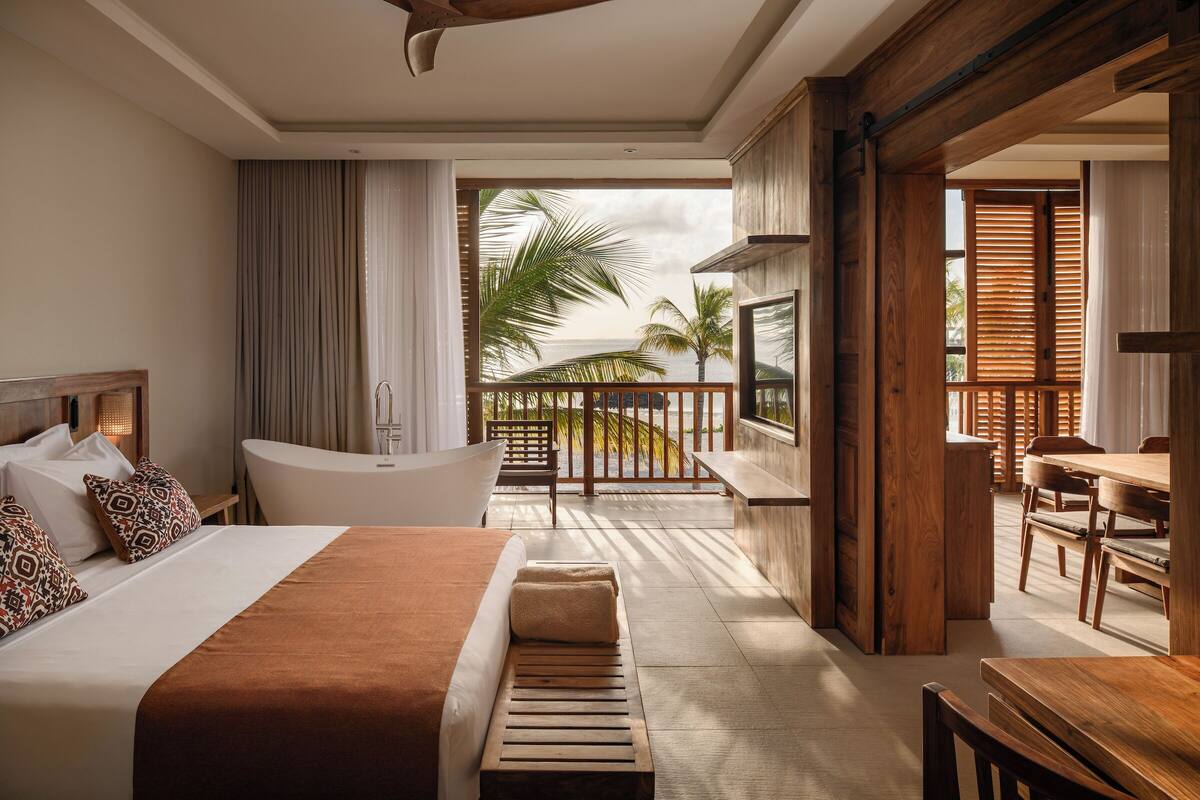 A hotel room with a large bed, a bathtub, a balcony with wooden railings, and a dining area with wooden furniture.