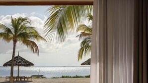 Une salle de bain moderne avec une baignoire sur pieds, une terrasse en bois et une vue sur une plage bordée de palmiers et abritée par un parasol en feuilles de palmier.