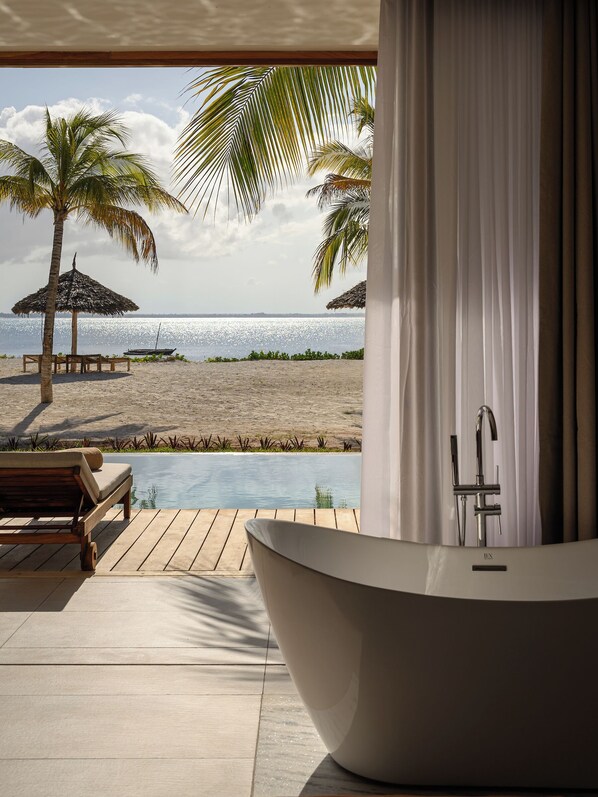 A modern bathroom with a freestanding bathtub, a wooden deck, and a view of a beach with palm trees and a thatched umbrella. - JAZ Elite Aurora (Michamvi)