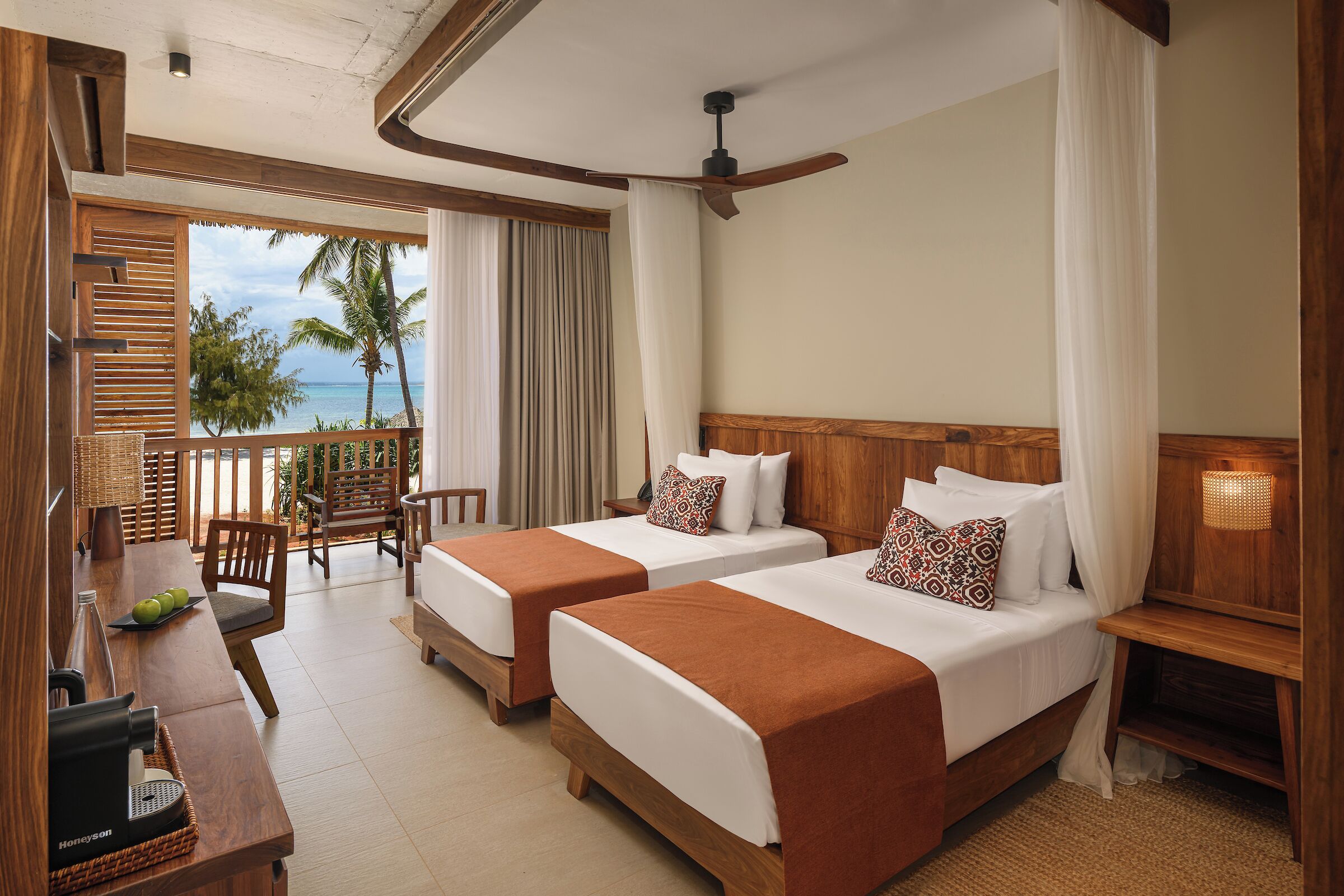 A hotel room with two beds, a balcony with a view of the beach, a dining area, and a ceiling fan.