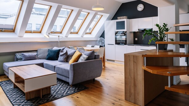 A modern apartment with a loft-style ceiling, a gray sofa with cushions, a wooden coffee table, a kitchen area with cabinets and a plant, and large skylights. - RentPlanet - Świętego Mikołaja (Wroclaw)