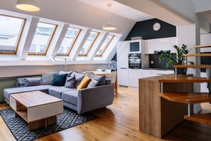 A modern apartment with a loft-style ceiling, a gray sofa with cushions, a wooden coffee table, a kitchen area with cabinets and a plant, and large skylights. - RentPlanet - Świętego Mikołaja (Wroclaw)