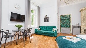 A modern hotel room with a green sofa, a dining area with chairs, a TV, a clock, and a large window. - Traveler's Best- Old Town Guesthouse (Kraków)