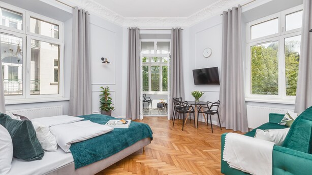 A modern hotel room with a wooden herringbone floor, a bed with a teal blanket, a green sofa, a dining area with a table and chairs, and large windows with curtains. - Traveler's Best- Old Town Guesthouse (Kraków)