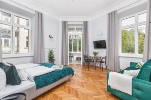 A modern hotel room with a wooden herringbone floor, a bed with a teal blanket, a green sofa, a dining area with a table and chairs, and large windows with curtains. - Traveler's Best- Old Town Guesthouse (Kraków)