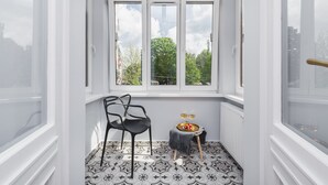 A room with a large window, a black chair, a small table with a tray of food and two glasses of wine, and patterned floor tiles. - Traveler's Best- Old Town Guesthouse (Kraków)