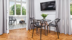 A modern room with a large window, a flat-screen TV, a round table with chairs, and a small side table with a vase of flowers. - Traveler's Best- Old Town Guesthouse (Kraków)