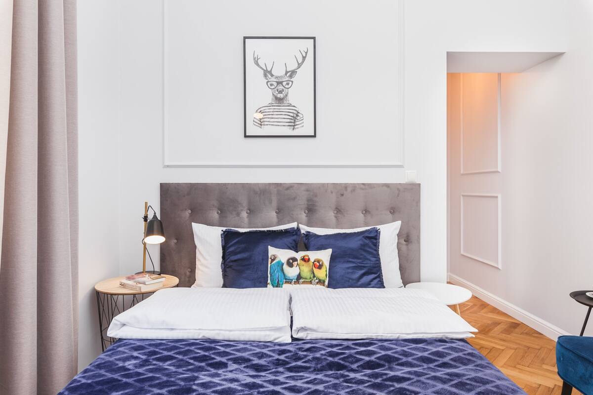 A neatly arranged bedroom with a large bed, a headboard, a bedside table with a lamp, and a framed picture on the wall.