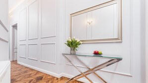 A hallway with white paneled walls, a glass console table with a vase of flowers, and a mirror with a gold frame. - Traveler's Best- Old Town Guesthouse (Kraków)