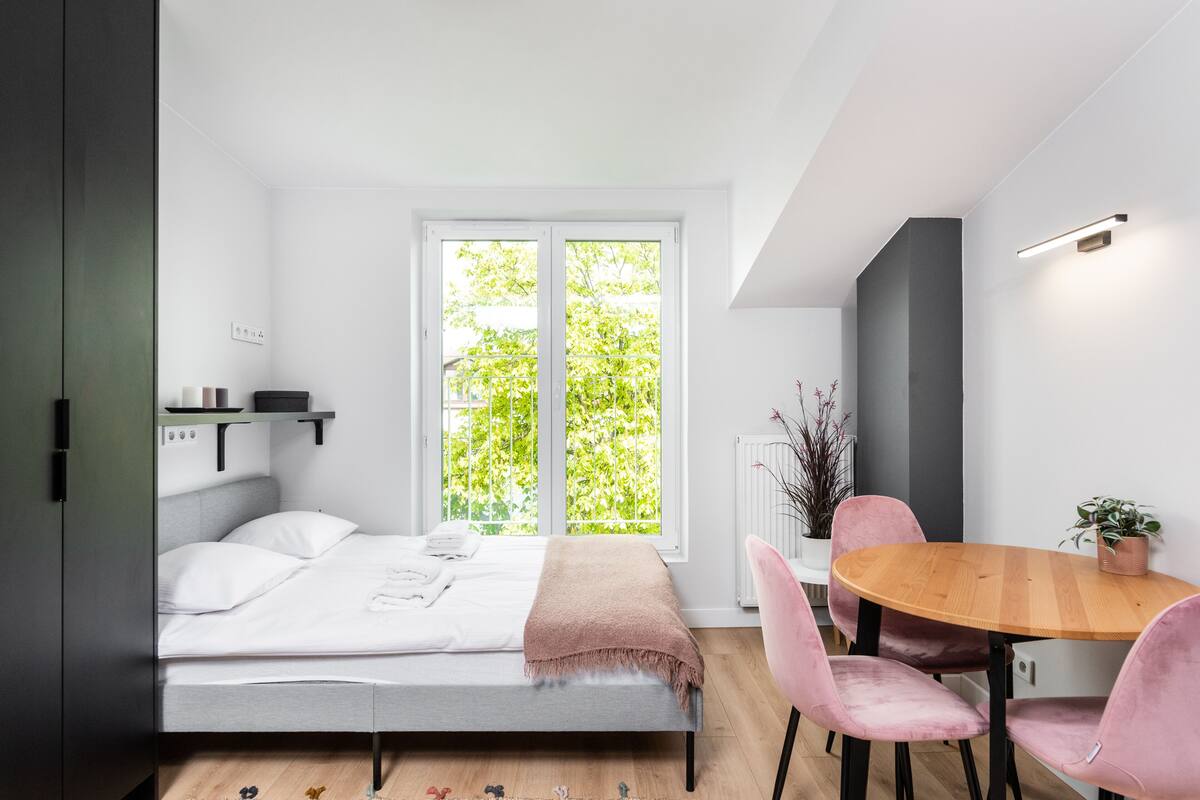 A modern bedroom with a bed, a window, a dining table with chairs, and a small plant.