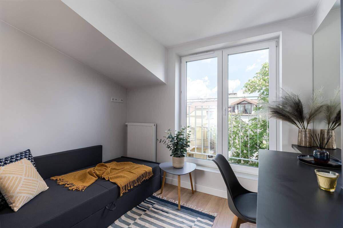 A modern living room with a dark sofa, a small round table, and a chair. There is a window with a view of buildings and greenery outside.