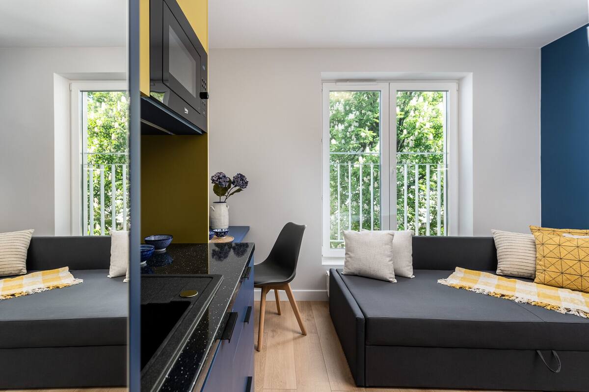 A modern living room with two black sofas, a small dining table, and a TV mounted on a yellow wall.