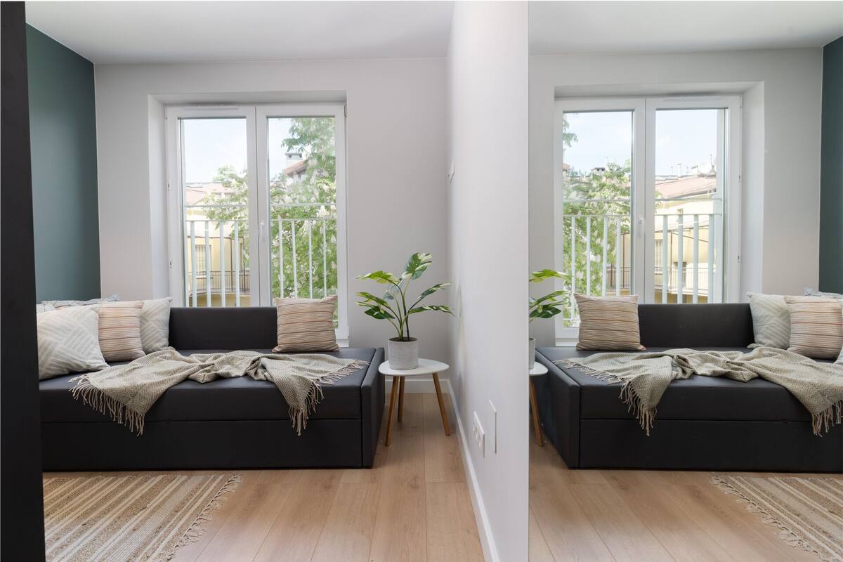 A modern living room with a black sofa, a small round table with a plant, and a view of a balcony with greenery.