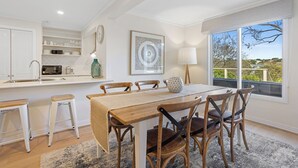Une cuisine moderne avec des meubles blancs, un coin repas doté d’une table et de chaises en bois, et une grande fenêtre offrant une vue sur l’extérieur.