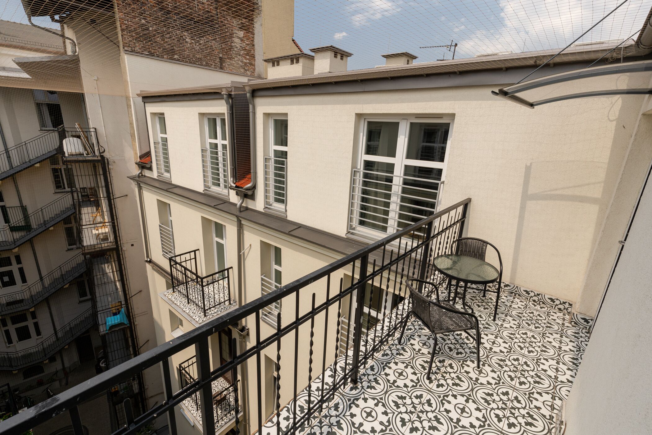 A multi-story building with a balcony featuring a patterned floor, a metal railing, and a table with chairs.