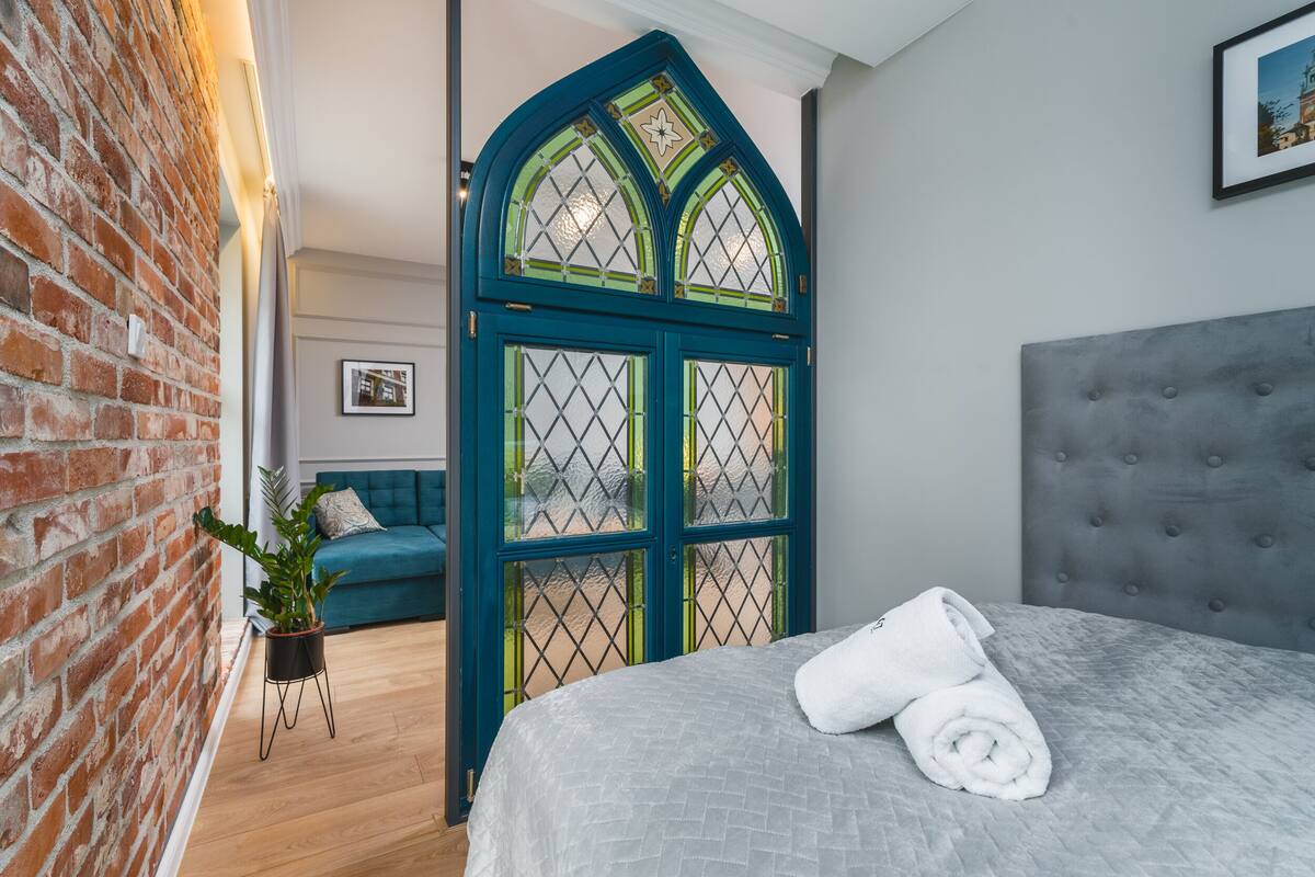 Un dormitorio con una cama, cabecera gris, puerta azul con paneles de vidrio, pared de ladrillo y una planta en maceta.