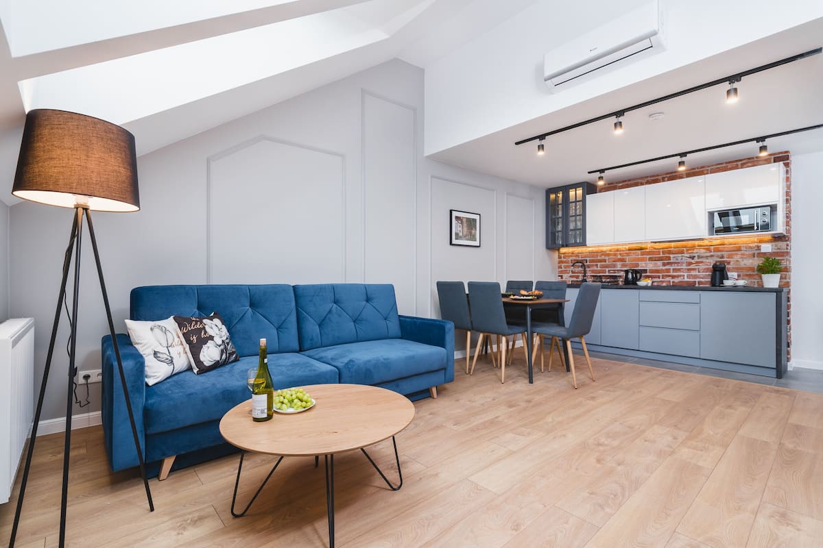 Una sala de estar moderna con un sofá azul, una mesa de centro redonda y una lámpara de pie. Hay un comedor con una mesa y sillas. La cocina tiene un salpicadero de ladrillo y electrodomésticos modernos.