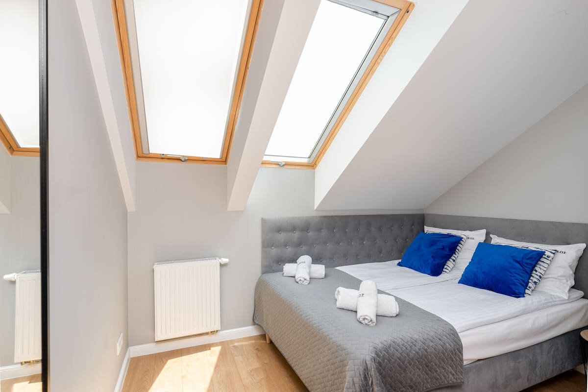Un dormitorio con techo inclinado, tragaluces, una cama gris con almohadas azules y un radiador blanco.