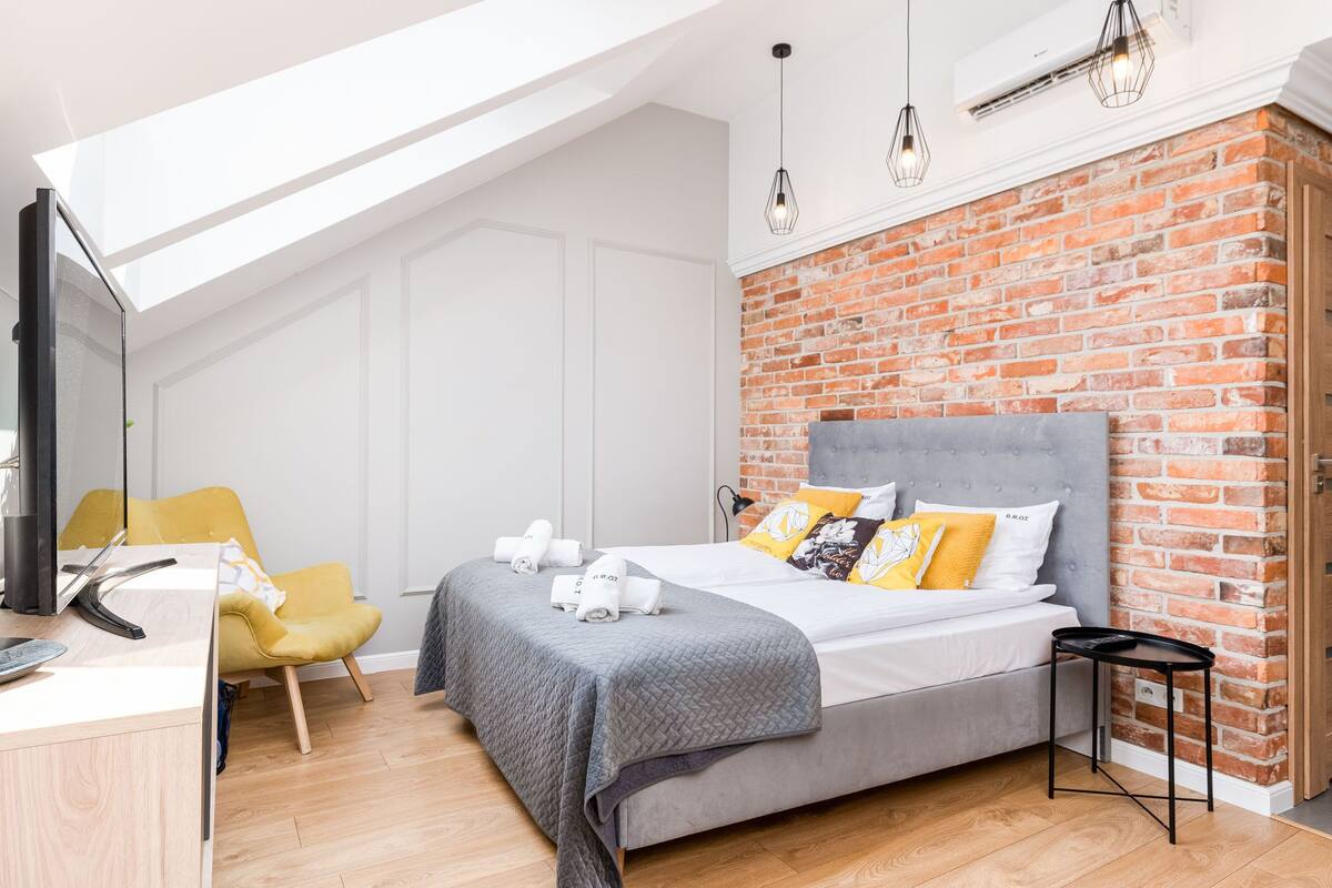 Un dormitorio moderno con pared de ladrillo, cama gris, almohadas amarillas y blancas, una mesita de noche negra pequeña y una silla amarilla.
