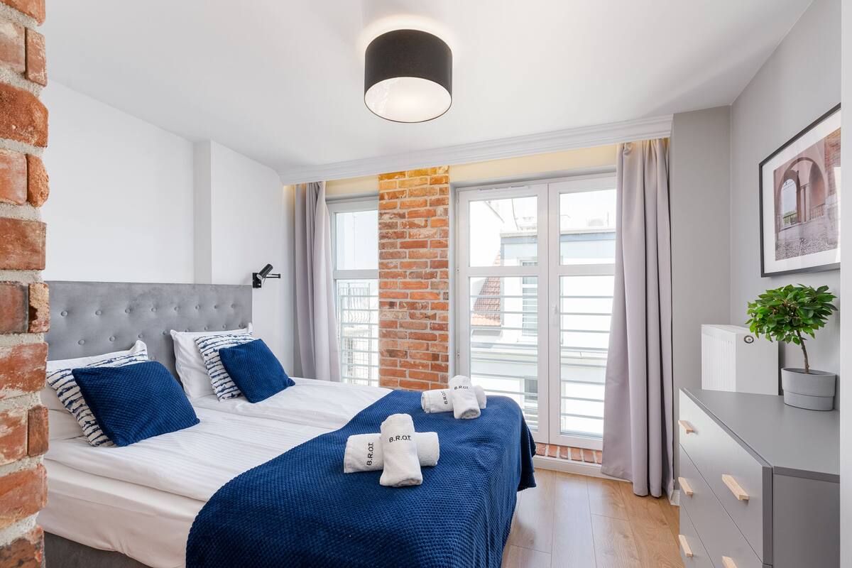 Habitación de hotel moderna con una cama grande, un muro de ladrillo decorativo, un cabecero gris y vistas a la ciudad a través de amplias ventanas.