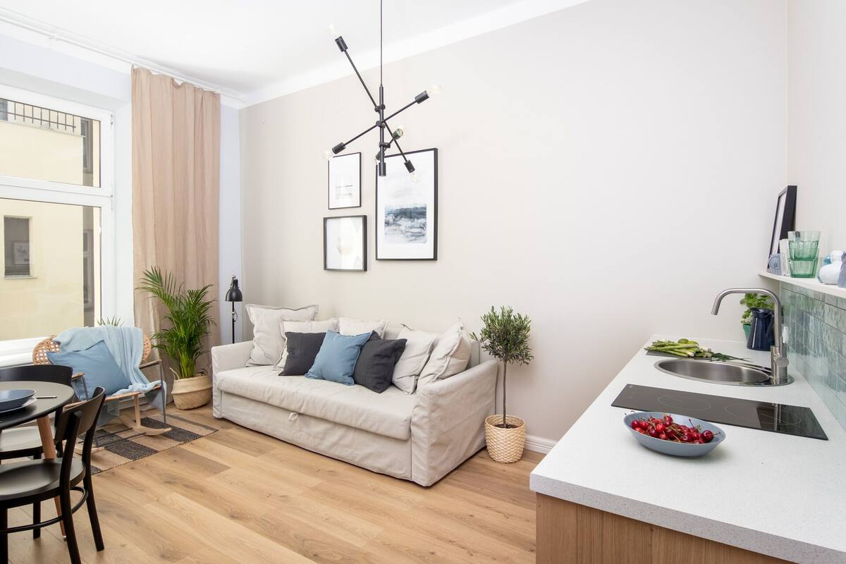 Una sala de estar moderna con un sofá, un comedor con sillas, una cocina con un fregadero y un cuenco de cerezas, y una ventana con cortinas.