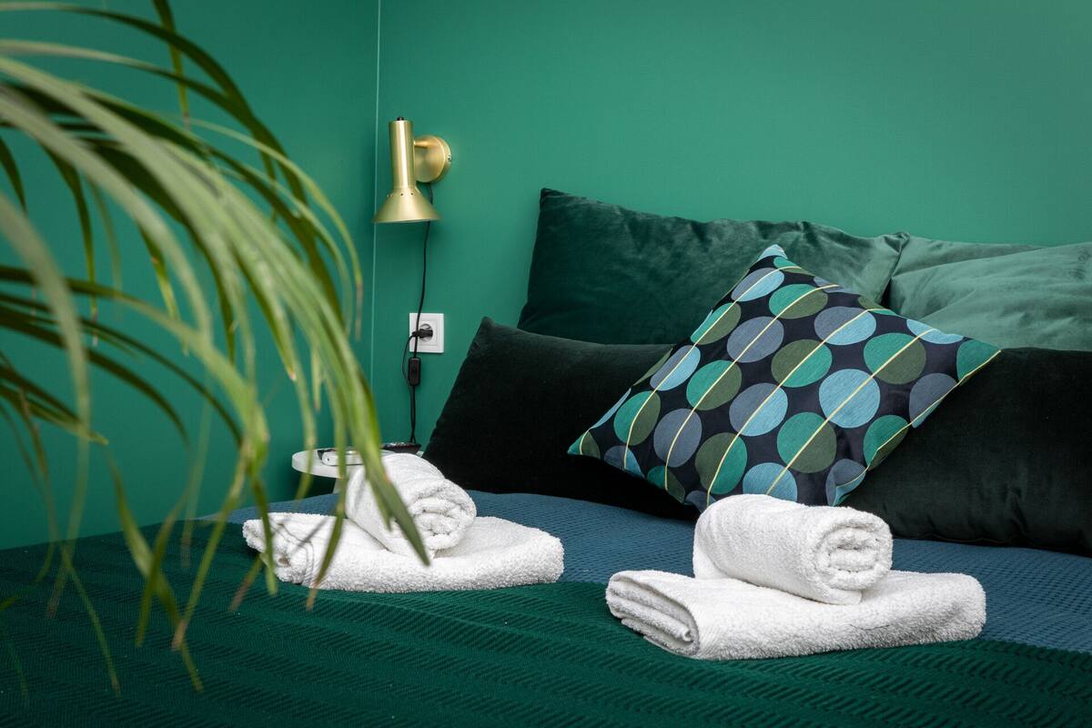 Una cama bien hecha con ropa de cama verde oscuro, una almohada estampada y dos toallas blancas enrolladas.