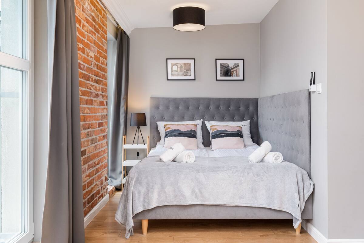 Un dormitorio moderno con una cama tapizada en gris, una pared de ladrillo y un ventanal grande.