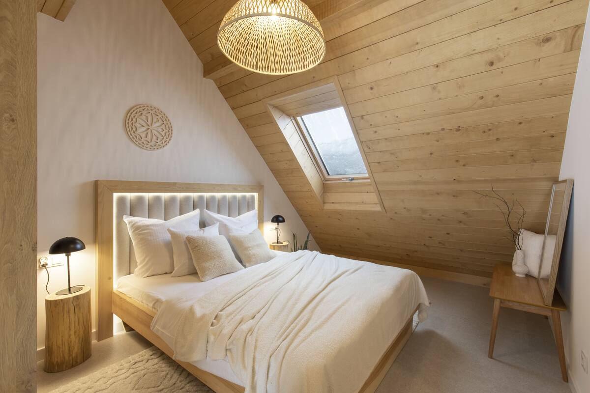 Une chambre avec un plafond en pente, un lit avec une literie blanche, un élément de décoration murale rond, une lampe de chevet et une petite table avec un vase.