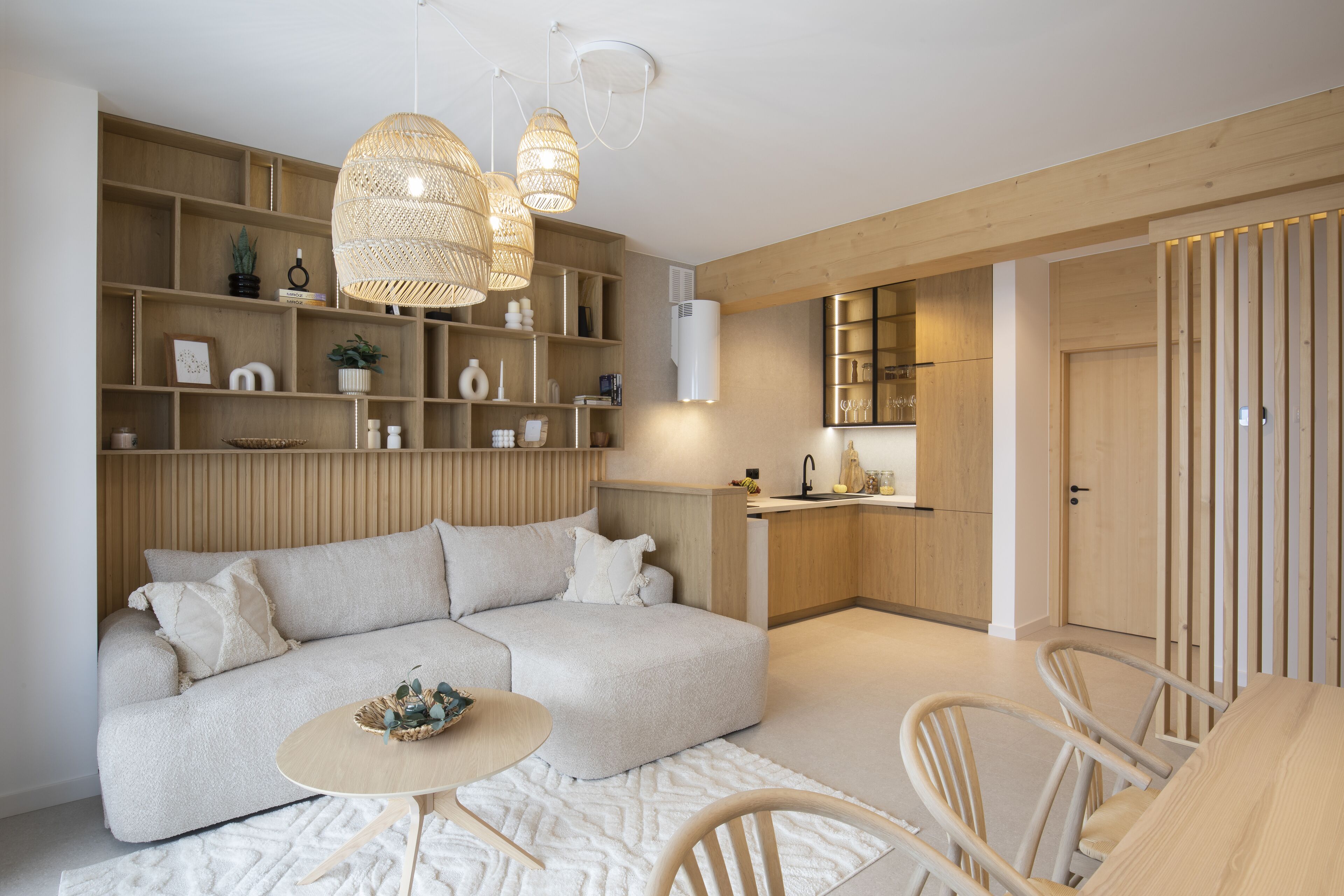 A modern living room with a sofa, dining table, and chairs. There are shelves with decorative items and a kitchen area in the background.
