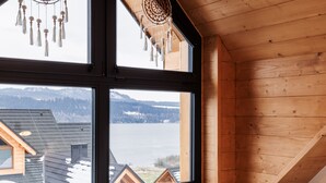 Une pièce chaleureuse avec un plafond en bois, une grande fenêtre donnant sur un lac et des bâtiments, et un lit bien fait, recouvert d’une couverture blanche.