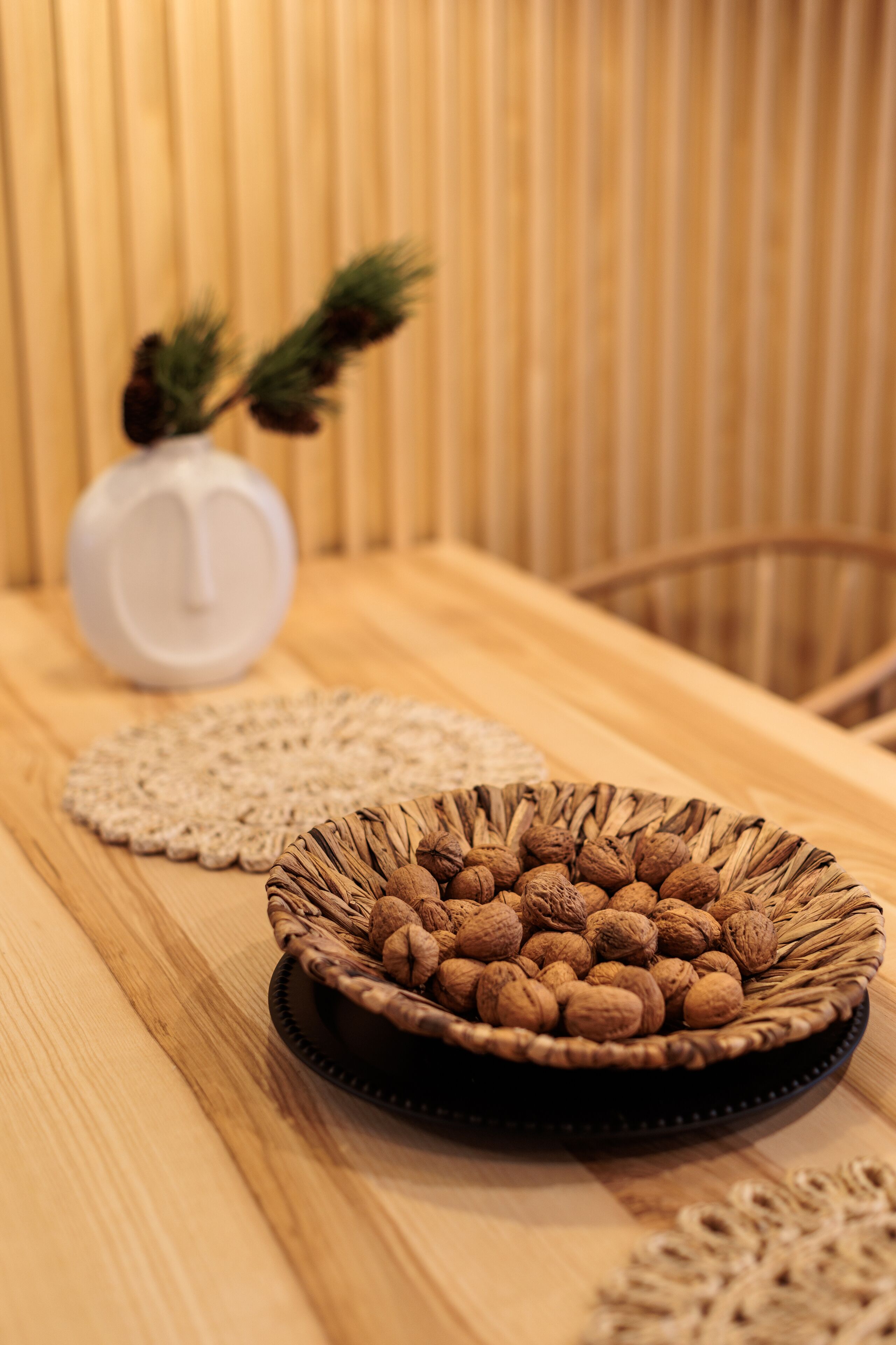 A wooden table with a woven bowl of nuts, a crocheted doily, and a white vase with pinecones.