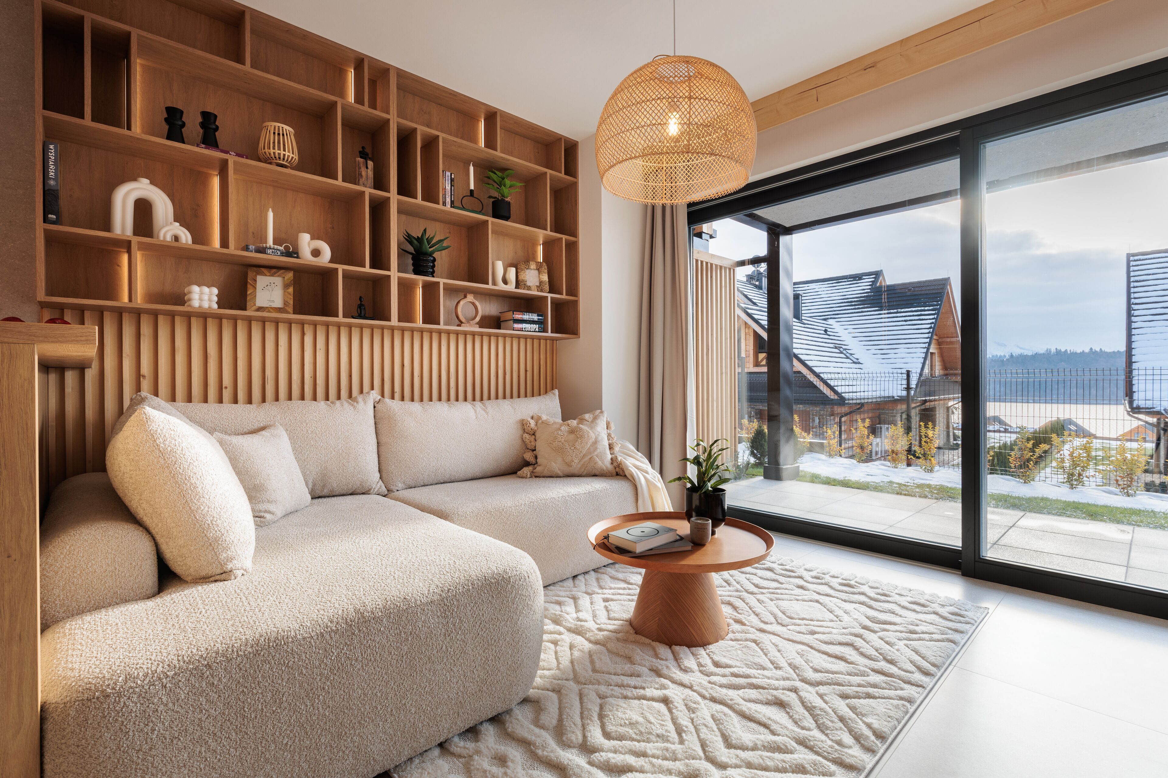Un salon moderne avec un canapé d’angle, une étagère en bois et une grande fenêtre donnant sur une maison.