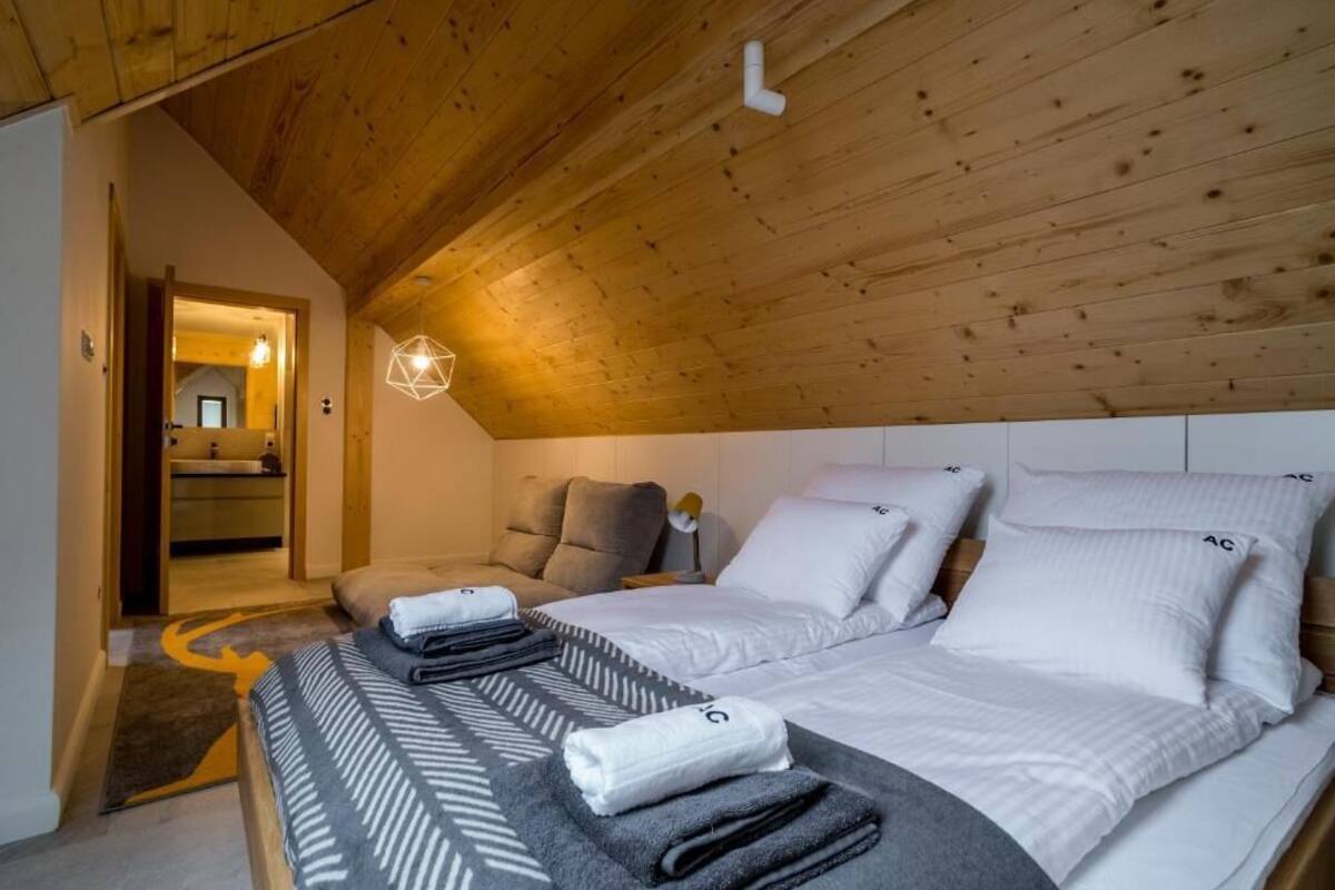 Une chambre confortable avec un plafond en pente, deux lits avec du linge de lit blanc, une couverture à motifs gris et blanc, et une tête de lit en bois.