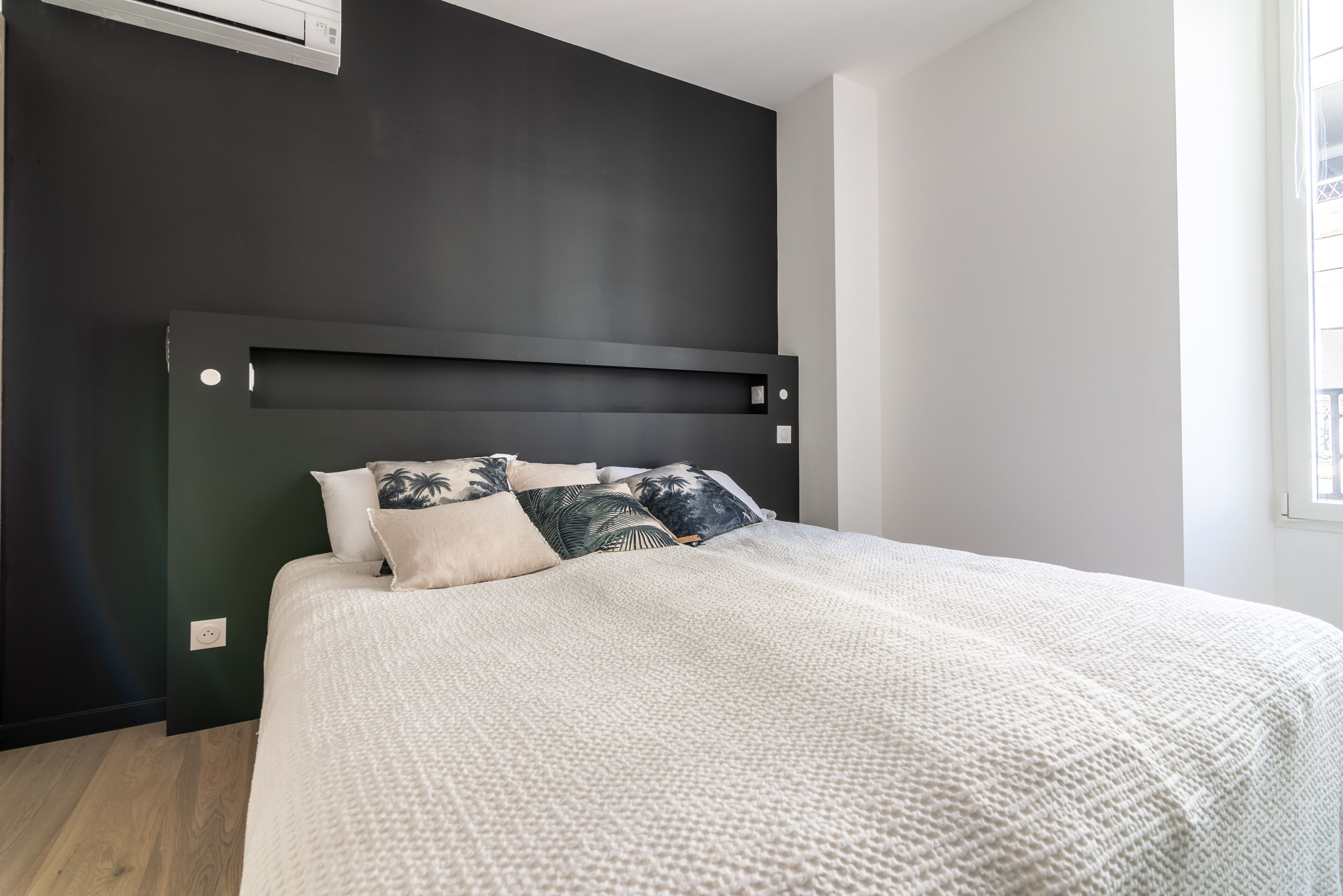 A modern bedroom with a dark headboard, a white bedspread, and tropical-themed pillows.