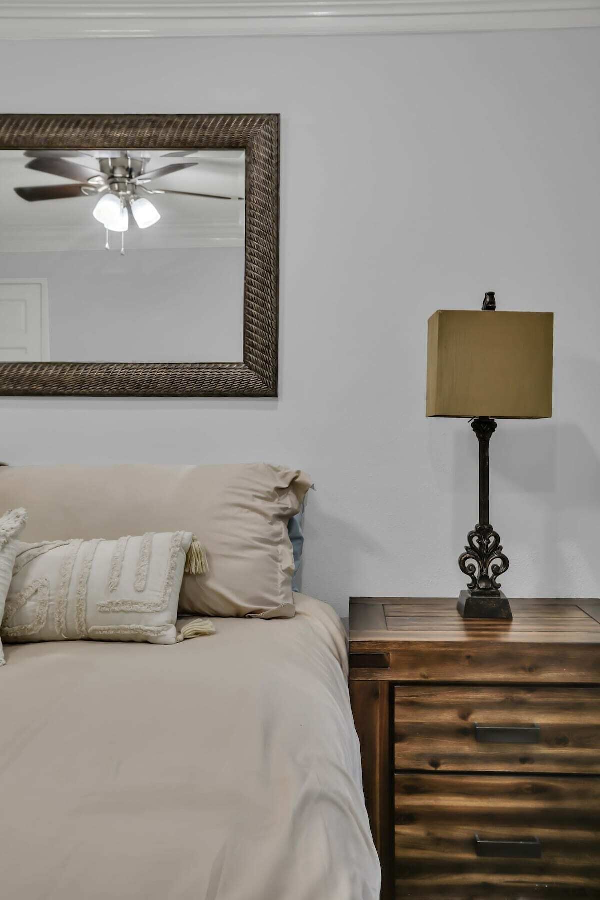 A bedroom with a bed, a ceiling fan, a bedside table with a lamp, and a mirror on the wall.