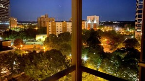 Vista nocturna de una ciudad desde una ventana, con edificios iluminados, árboles y un cielo despejado.