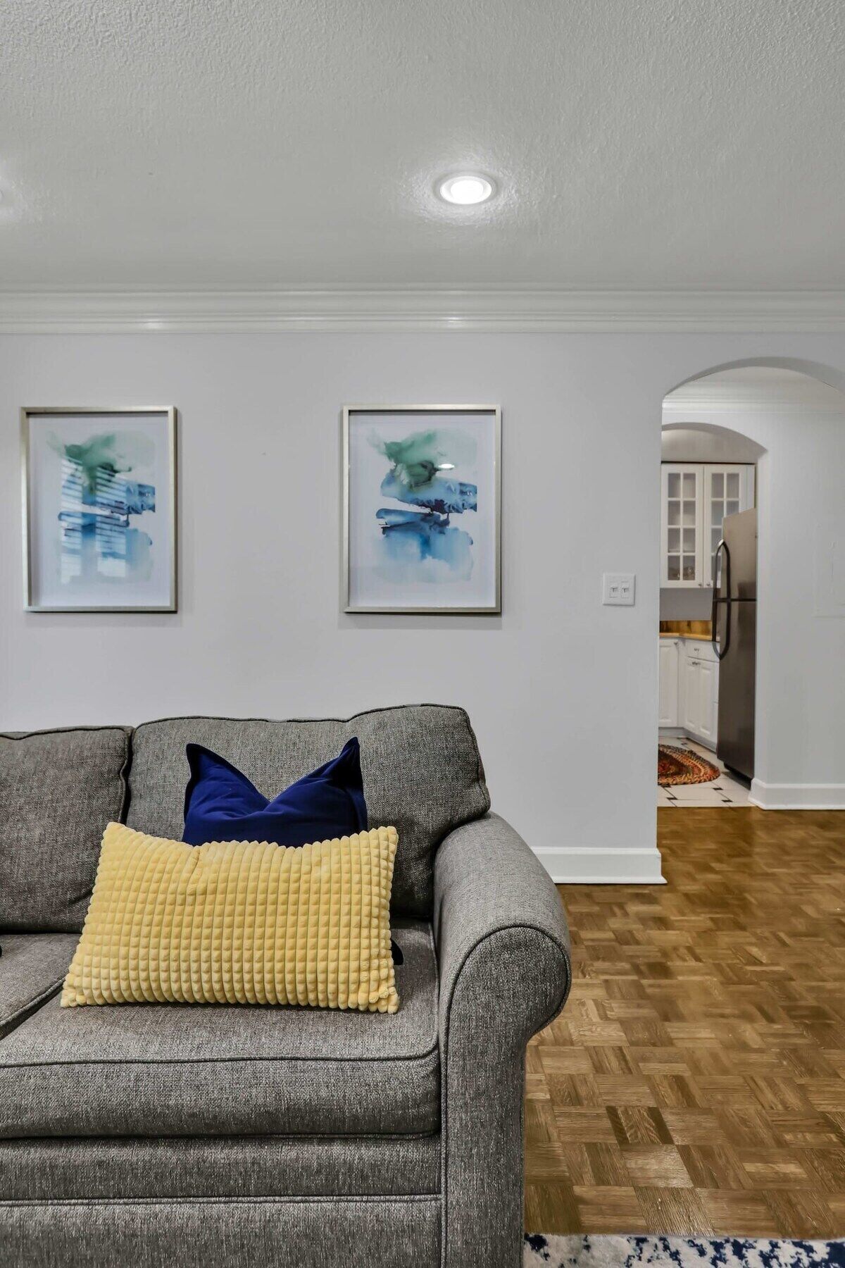 Un salon avec un canapé gris, deux tableaux abstraits et une cuisine visible à travers une arche.