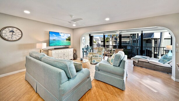 A spacious living room with a large flat-screen TV, a clock, and a balcony with outdoor seating.
