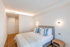 A modern bedroom with a large bed, a bedside table, a wall-mounted light, and a door leading to another room. - Renovato Prestige (Porto)