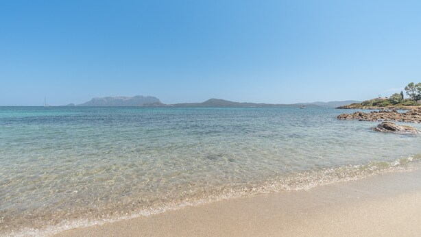 Ein Strand mit klarem Wasser, felsiger Küste und Bergen im Hintergrund.