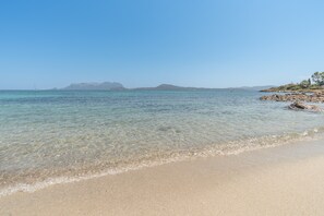 Ein Strand mit klarem Wasser, felsiger Küste und Bergen im Hintergrund.