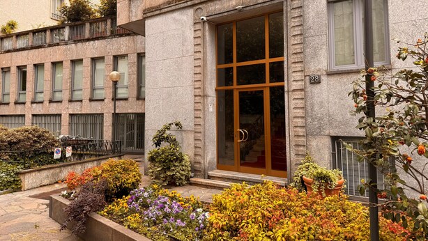 Un bâtiment doté d’une grande entrée en verre, d’une façade en pierre et d’un petit jardin où poussent différentes espèces de plantes.