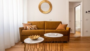 Un salon avec un canapé marron, un grand miroir rond doré et deux tables rondes aux pieds dorés.
