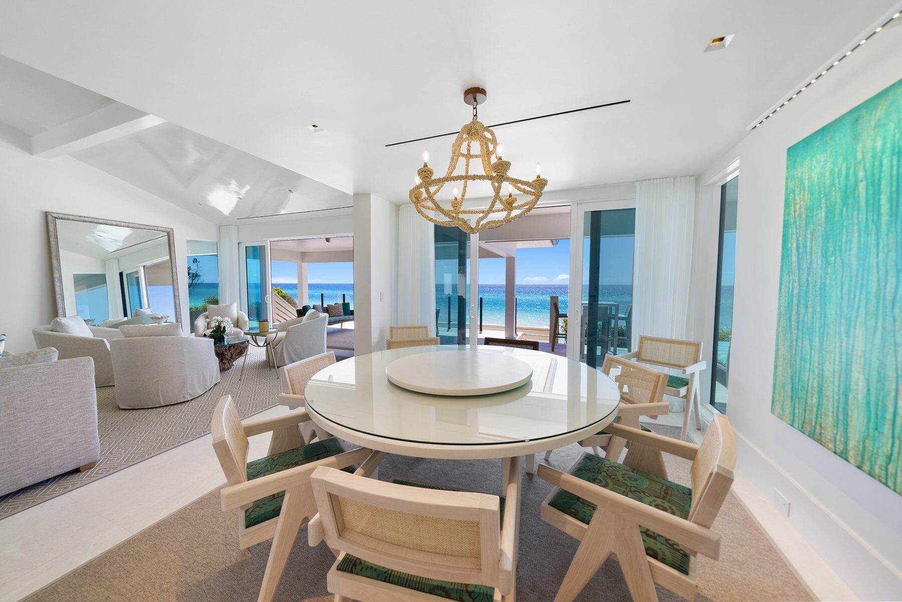 A modern dining area with a round glass table and wooden chairs, a chandelier, and a view of the ocean through large windows.