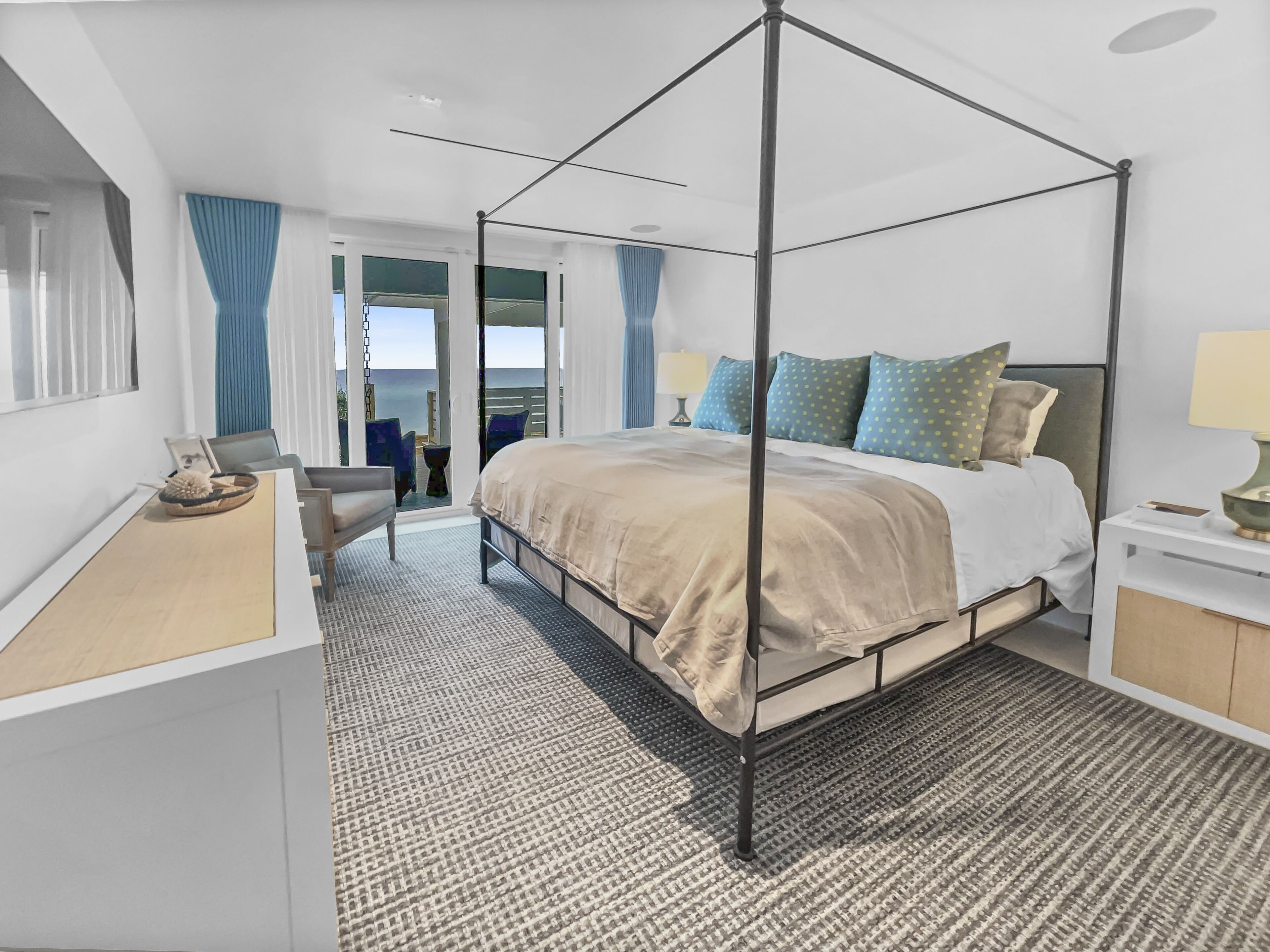 A modern bedroom with a four-poster bed, a desk with a chair, and a view of the ocean through large windows.