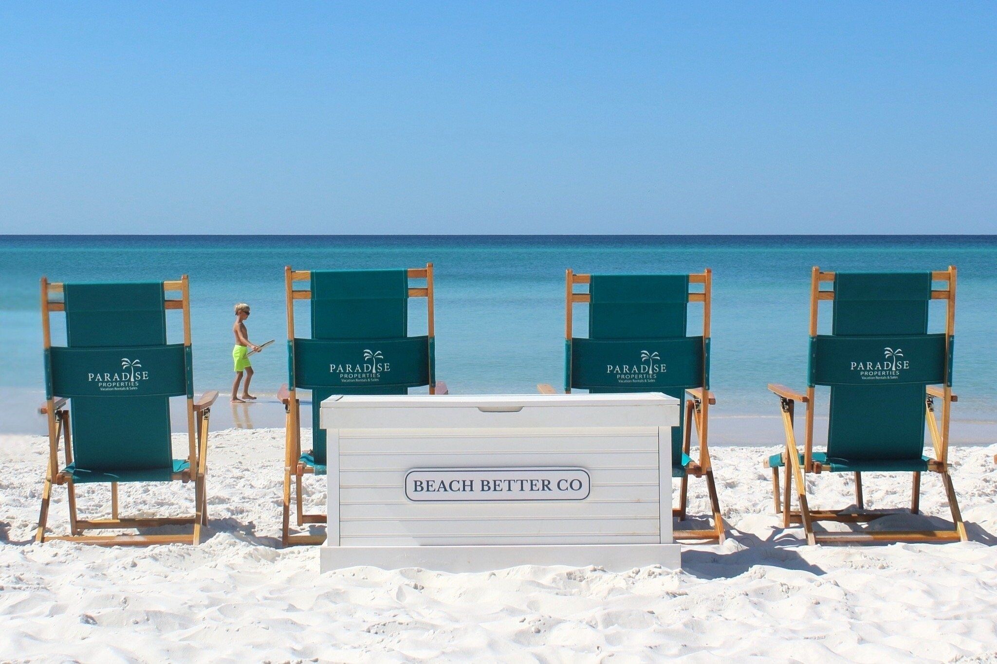 Une plage avec quatre chaises longues vertes portant l’inscription « Paradise Properties » et une glacière portant l’inscription « Beach Better Co. ».