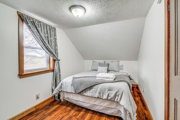 A bedroom with a sloped ceiling, a window with curtains, a bed with a gray blanket, and wooden flooring. - Cozy New York Farmhouse w/ Porch, Grill & Fire Pit (Geneva)