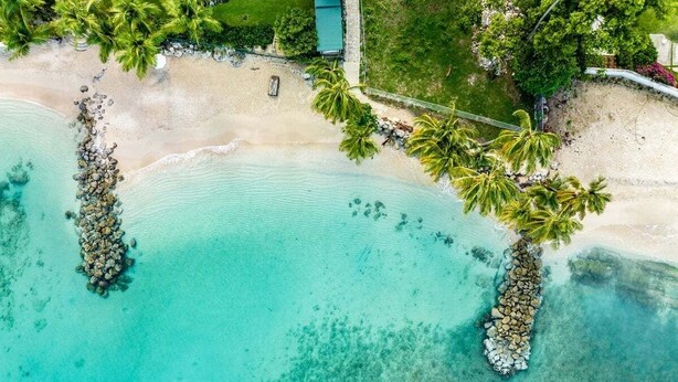 An aerial view of a beach with clear turquoise waters, palm trees, and a small building.